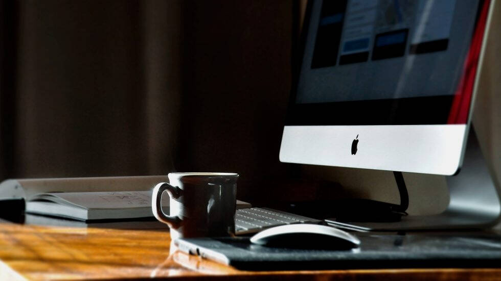 A desktop workspace featuring an Apple iMac on the right, partially visible with a white keyboard and Magic Mouse on a wooden desk, next to a dark coffee mug. Soft lighting highlights the glossy surface of the monitor and the warm wood grain of the desk.